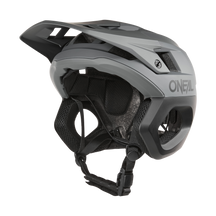 Der O'NEAL TRAPPER Helm SOLID in Grau/Schwarz besticht durch sein kantiges Design mit Belüftungsschlitzen und einem Fidlock® Magnetverschluss. Von der Seite zeigt er einen verstellbaren Kinnriemen und eine Innenpolsterung mit prominentem O'NEAL-Logo.