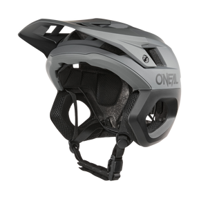 Der O'NEAL TRAPPER Helm SOLID in Grau/Schwarz besticht durch sein kantiges Design mit Belüftungsschlitzen und einem Fidlock® Magnetverschluss. Von der Seite zeigt er einen verstellbaren Kinnriemen und eine Innenpolsterung mit prominentem O'NEAL-Logo.