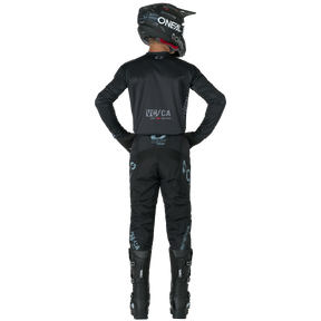 Eine Person in schwarzer O'NEAL ELEMENT-Hose, Helm, Handschuhen und Stiefeln steht mit den Händen auf den Hüften und schaut weg. Die Logos der Marke O'NEAL sind auf dem Anzug und dem Helm sichtbar.