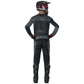 Eine Person in schwarzer und grauer Motocross-Ausrüstung steht mit den Händen in den Hüften, trägt ein O'NEAL-Trikot, hohe schwarze Stiefel und O'NEAL ELEMENT Pants RACEWEAR.