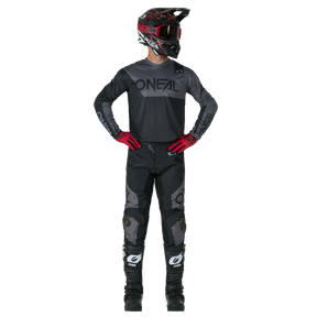 Eine Person in einem kompletten Motocross-Outfit, bestehend aus schwarzer O'NEAL ELEMENT Pants RACEWEAR, einem schwarzen O'NEAL-Helm, Trikot, roten Handschuhen und schwarzen Stiefeln, steht mit den Händen in den Hüften vor einem unifarbenen Hintergrund.