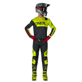 Eine Person steht mit den Händen in den Hüften und blickt zur Seite. Sie trägt O'NEAL ELEMENT Pants RACEWEAR in Schwarz und Neongelb, gepaart mit passendem O'NEAL Helm, Handschuhen, Trikot und Stiefeln.