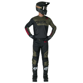 Eine Person in kompletter Motocross-Kleidung, einschließlich schwarzem Helm mit Brille, dunklem O'NEAL-Trikot, O'NEAL ELEMENT Pants RACEWEAR, Handschuhen und schwarzen O'NEAL-Stiefeln, steht mit den Händen auf den Hüften und schaut leicht zur Seite.