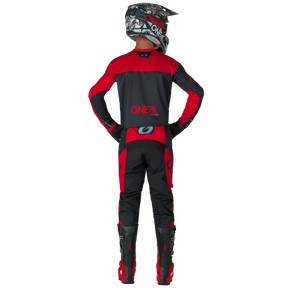 Ein Motocross-Fahrer wird von hinten gezeigt. Er steht mit den Händen in den Hüften und trägt O'NEAL ELEMENT Pants RACEWEAR in Schwarz und Rot sowie passende O'NEAL-Stiefel, Handschuhe und einen Helm.