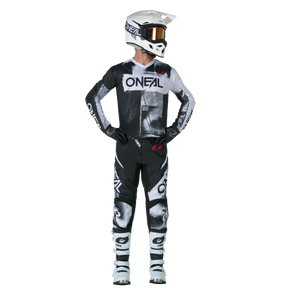 Ein Motocross-Fahrer steht aufrecht und blickt zur Seite. Er trägt den O'NEAL ELEMENT Pants ROLLER sowie einen passenden schwarz-weißen O'NEAL-Helm, eine Schutzbrille, Handschuhe und Stiefel.