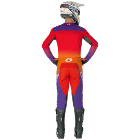 Eine Person in einem roten, orangefarbenen und lilafarbenen O'NEAL MAYHEM Hose HEXX-Motocross-Anzug mit schwarzen Stiefeln und einem weißen Helm steht mit den Händen in den Hüften. Das Outfit weist O'NEAL-Branding auf dem Helm, dem Rücken und den Beinen auf.
