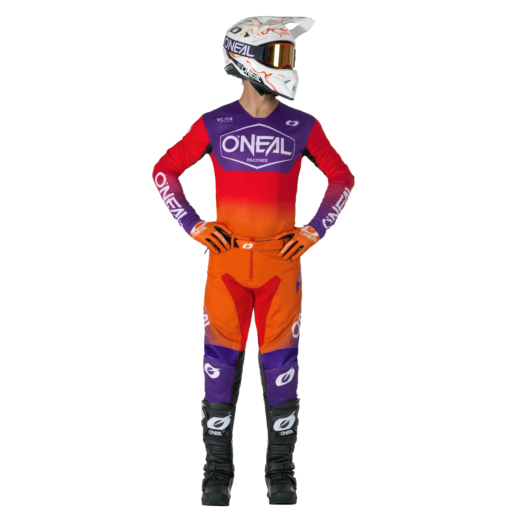 Ein Motocross-Fahrer mit weißem Helm, rotem und orangefarbenem O'NEAL-Trikot, O'NEAL MAYHEM Hose HEXX Hose mit lila Akzenten und schwarzen Stiefeln steht mit den Händen in den Hüften und schaut zur Seite.