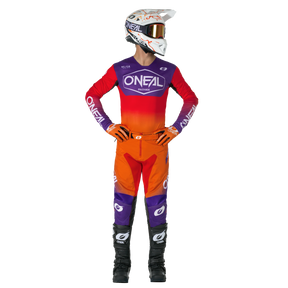 Ein Motocross-Fahrer mit weißem Helm, rotem und orangefarbenem O'NEAL-Trikot, O'NEAL MAYHEM Hose HEXX Hose mit lila Akzenten und schwarzen Stiefeln steht mit den Händen in den Hüften und schaut zur Seite.