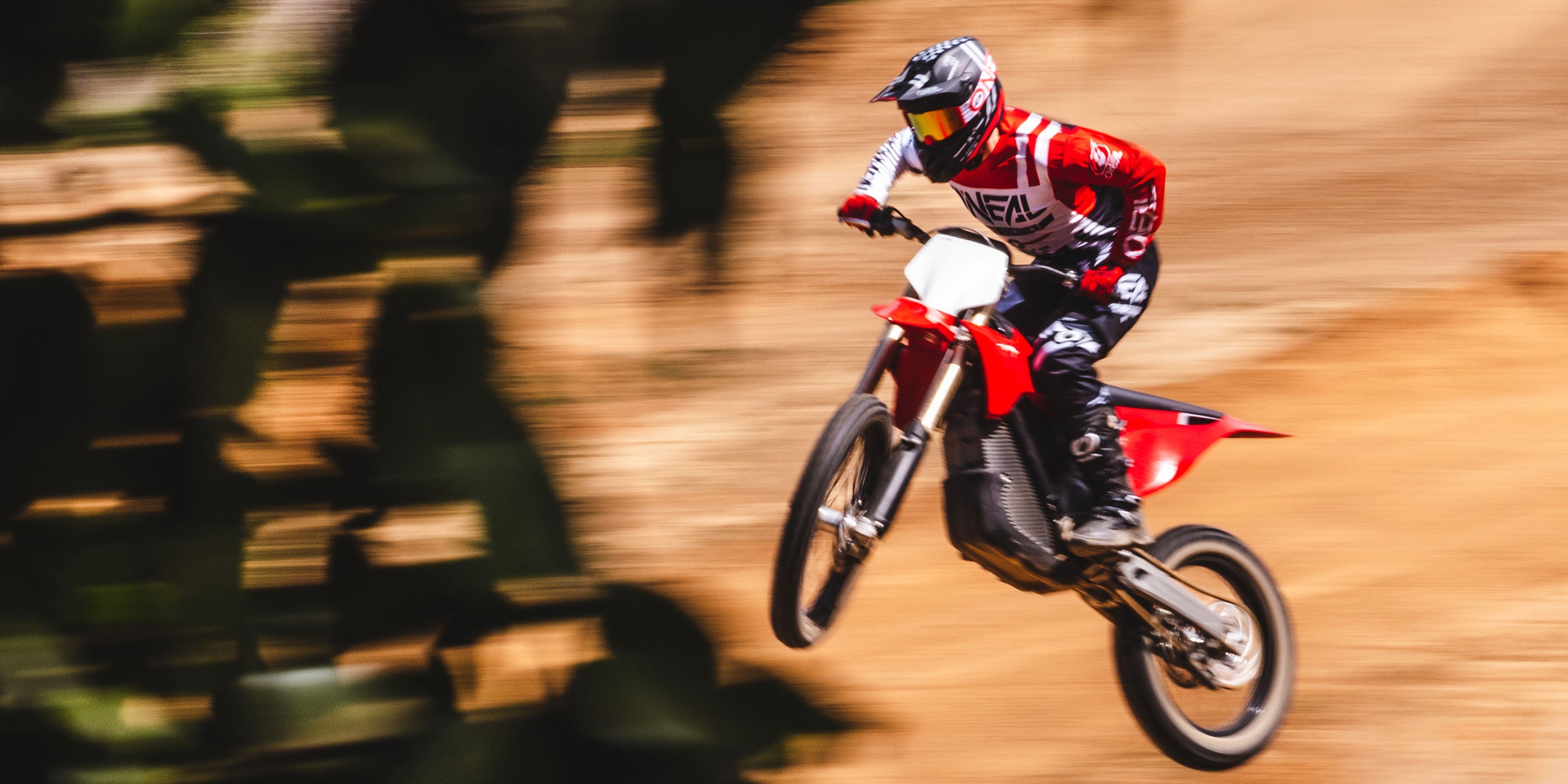 Ein Motocrossfahrer in rot-weißer Ausrüstung und mit Helm ist auf einem roten Dirtbike in der Luft und macht einen Sprung auf einer unbefestigten Strecke. Der Hintergrund ist verschwommen, was die Bewegung und Geschwindigkeit des Fahrers hervorhebt. Dunkles Laub ist teilweise zu sehen, das die linke Seite des Bildes einrahmt.