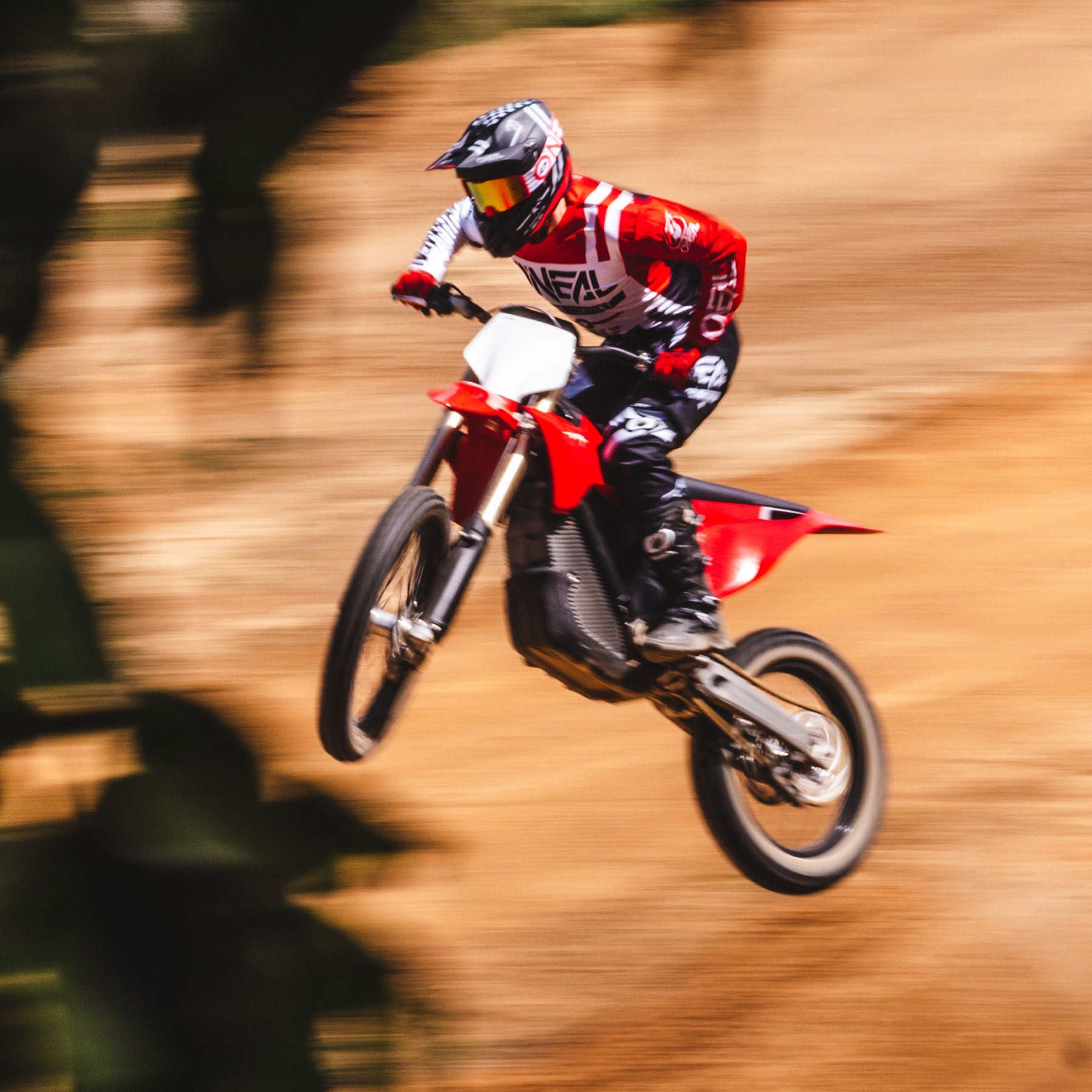 Ein Motocrossfahrer in rot-weißer Ausrüstung macht einen Sprung auf einer unbefestigten Strecke. Der Hintergrund erscheint verschwommen, was die Geschwindigkeit und Bewegung des Motorrads hervorhebt. Der Fahrer trägt einen Helm und eine Schutzbrille und blickt nach vorne.