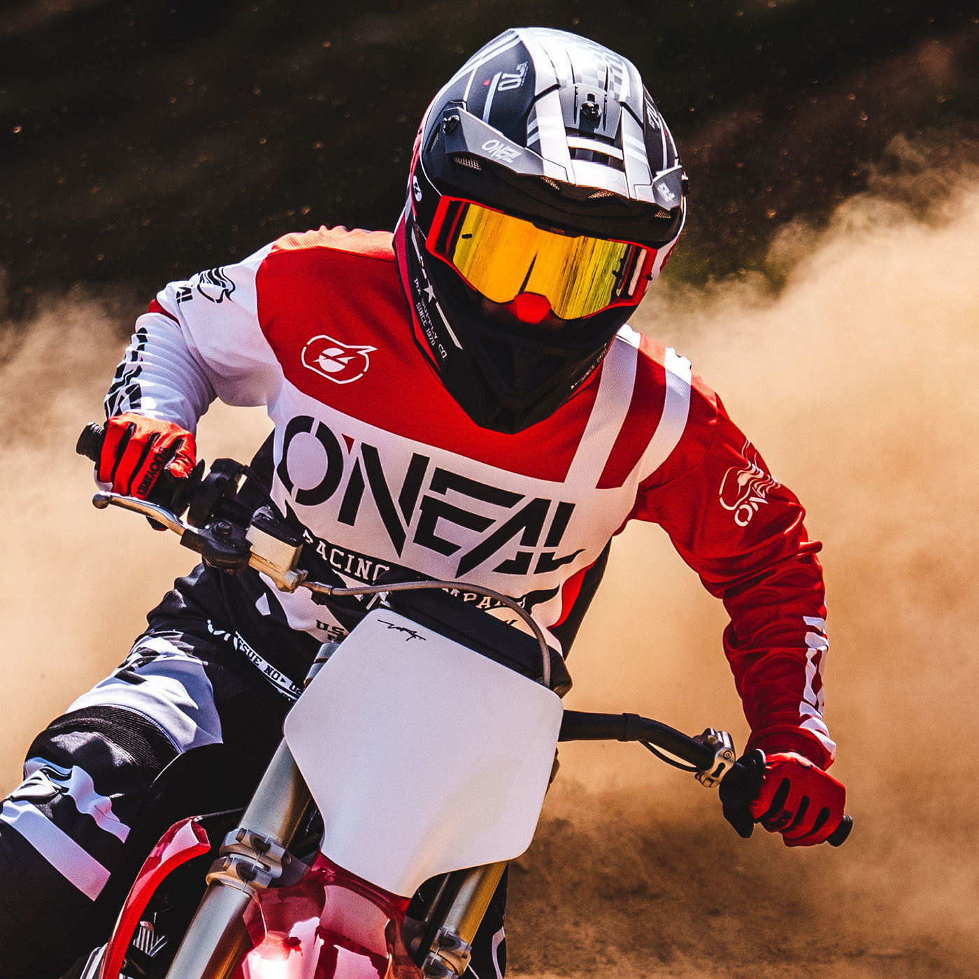 Ein Motocrossfahrer mit schwarzem Helm und gelbem Visier und rot-schwarz-weißer O'Neal-Rennausrüstung fährt mit einem Dirtbike durch eine Staubwolke auf einer Rennstrecke. Der Fahrer hält den Lenker fest umklammert, während er sich in eine Kurve lehnt.