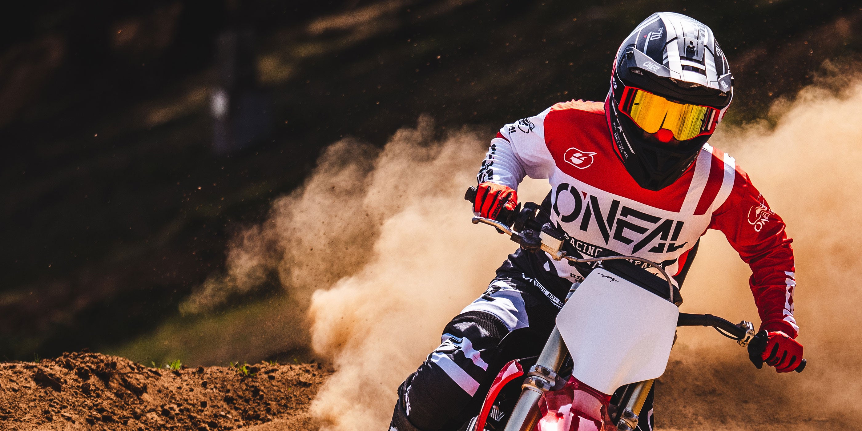 Eine Person in Schutzkleidung, darunter ein Helm mit getöntem Visier und ein Trikot mit der Aufschrift „ONEAL“, fährt mit einem Dirtbike über eine staubige Strecke und wirbelt dabei eine Staubwolke hinter sich auf. Das Bild fängt die intensive Action und Bewegung des Motocross ein.