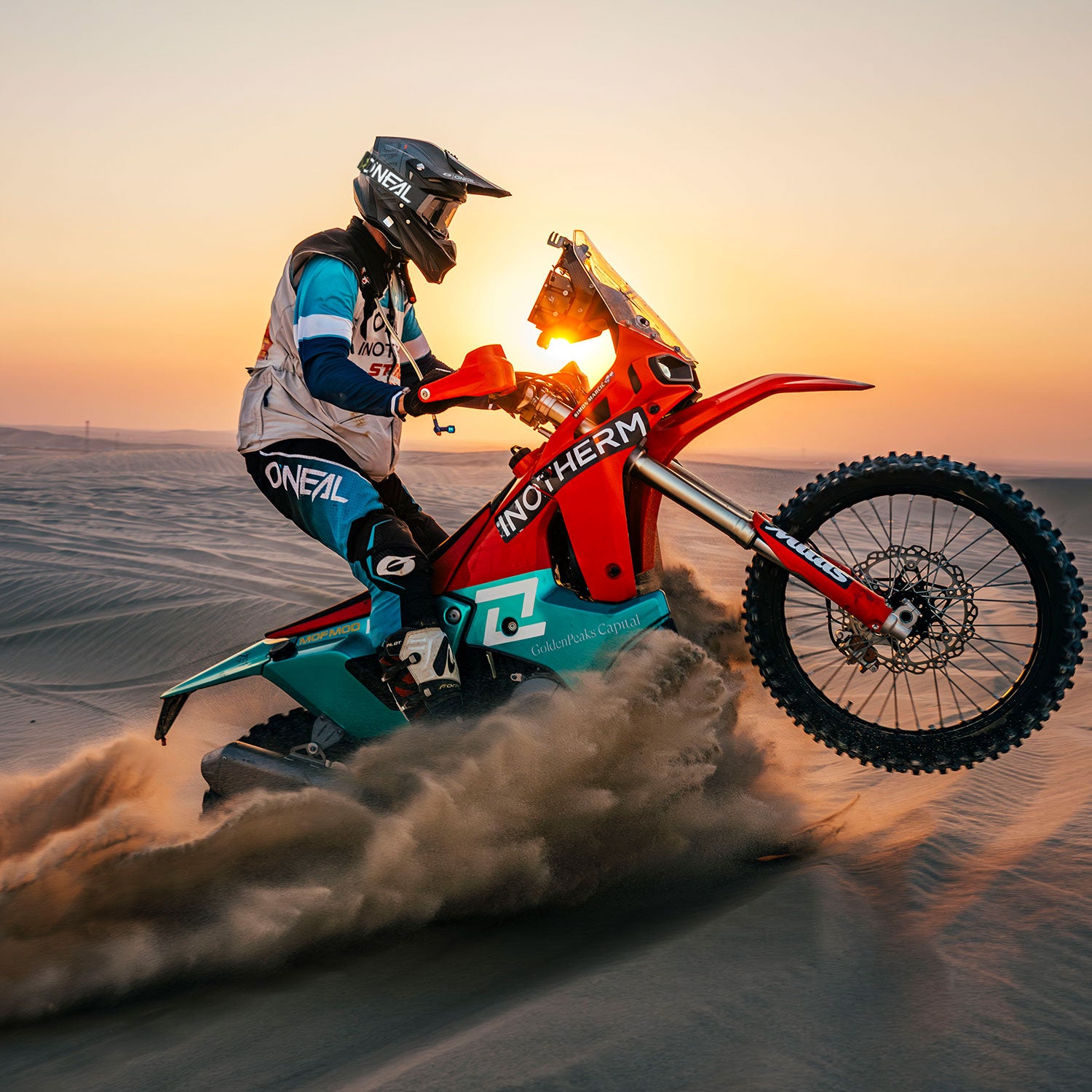 Ein Motocross-Fahrer in voller Montur vollführt einen Wheelie auf einem roten und blaugrünen Dirt Bike und wirbelt dabei Sand auf, während die Sonne über einer weiten Wüstenlandschaft untergeht.