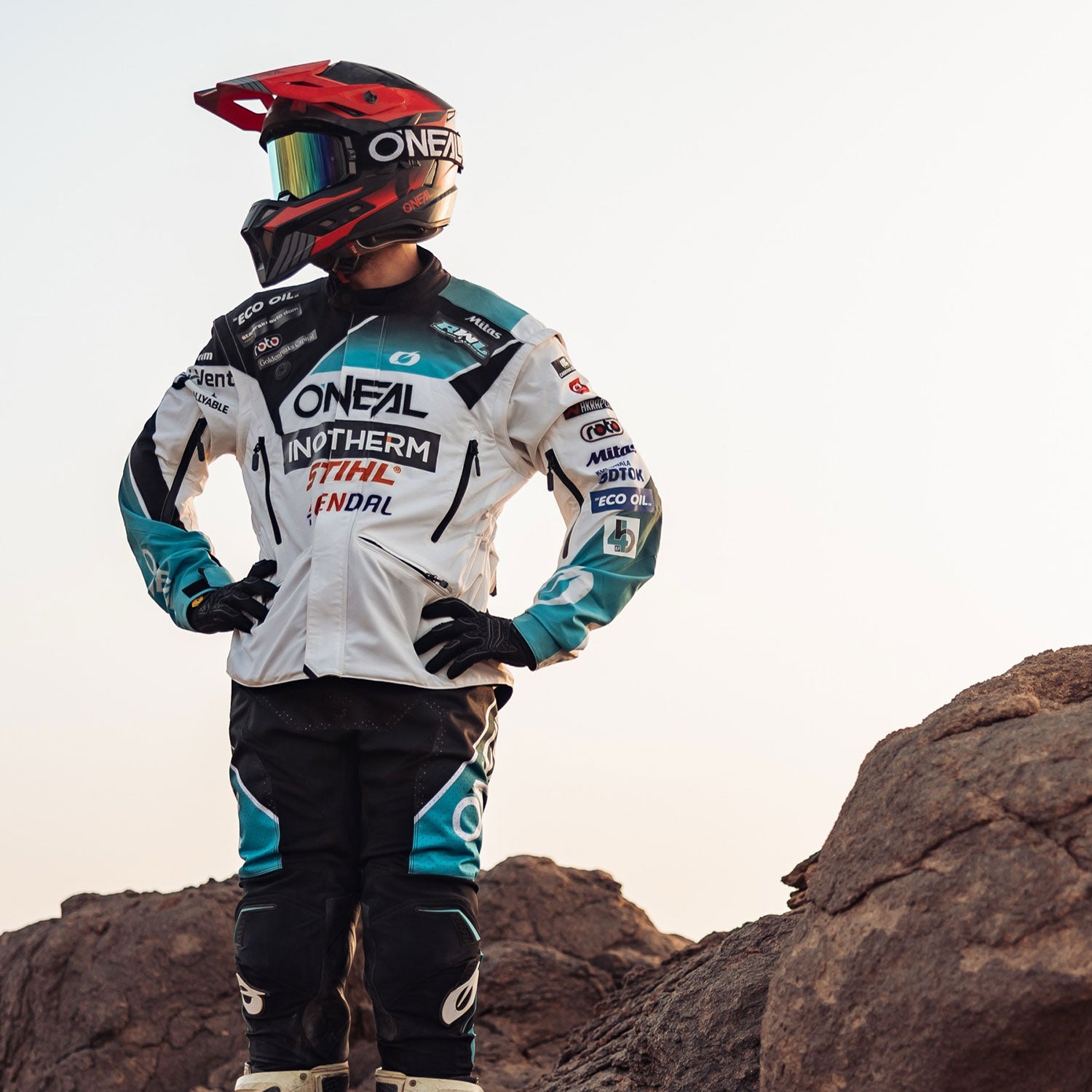 Eine Person in kompletter Motocross-Ausrüstung, einschließlich Helm, Handschuhen und Schutzausrüstung, steht auf einem felsigen Gelände, die Hände in die Hüften gestemmt, und blickt bei klarem Himmel zur Seite.