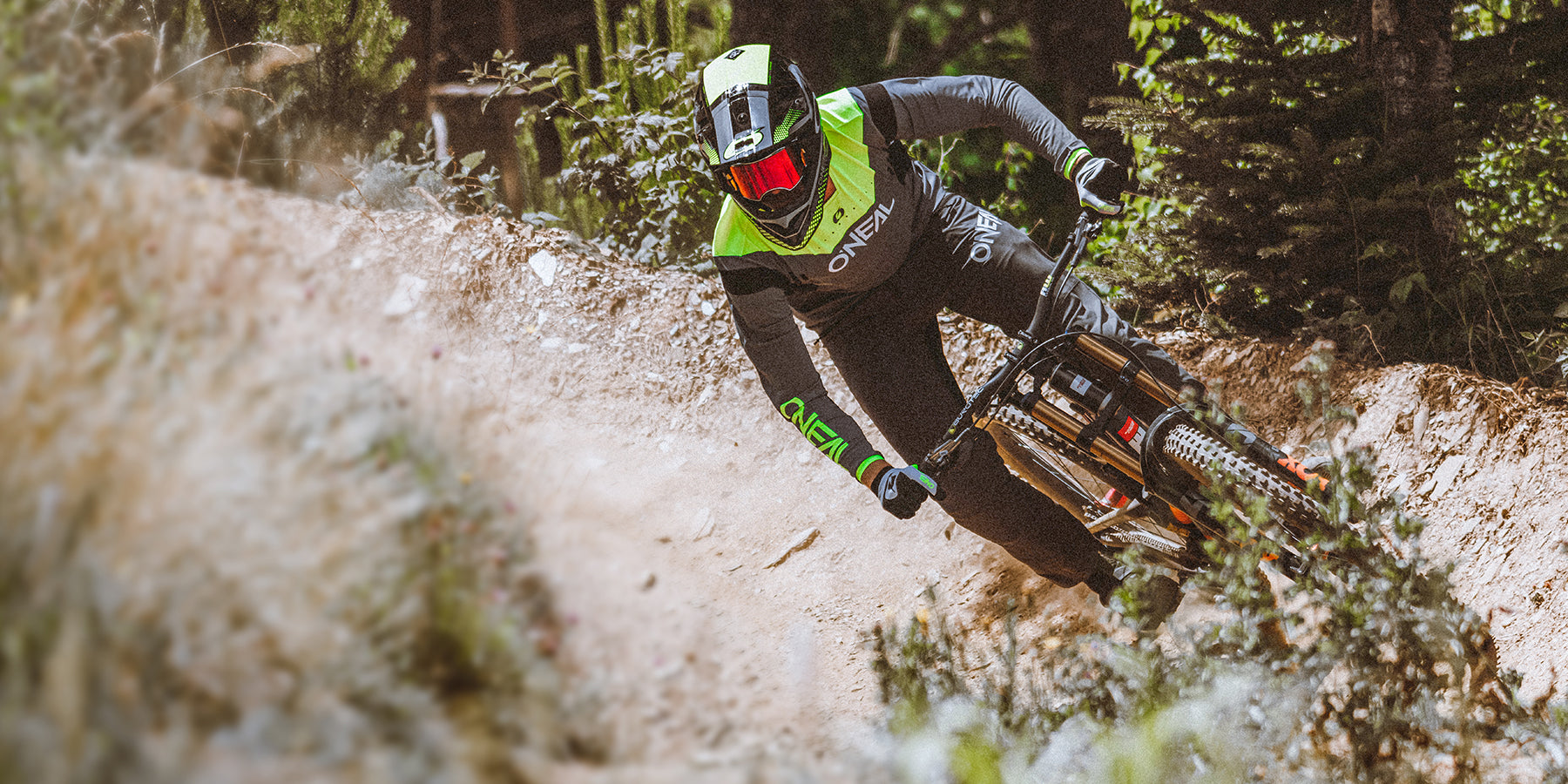 Ein Mountainbiker in voller Montur, einschließlich Helm und Schutzbrille, fährt einen Feldweg durch einen Wald hinunter. Der Biker legt sich in eine Kurve, im Hintergrund sind Grün und Bäume zu sehen. Die Szene fängt rasante Action und den Nervenkitzel des Downhill-Mountainbikings ein.