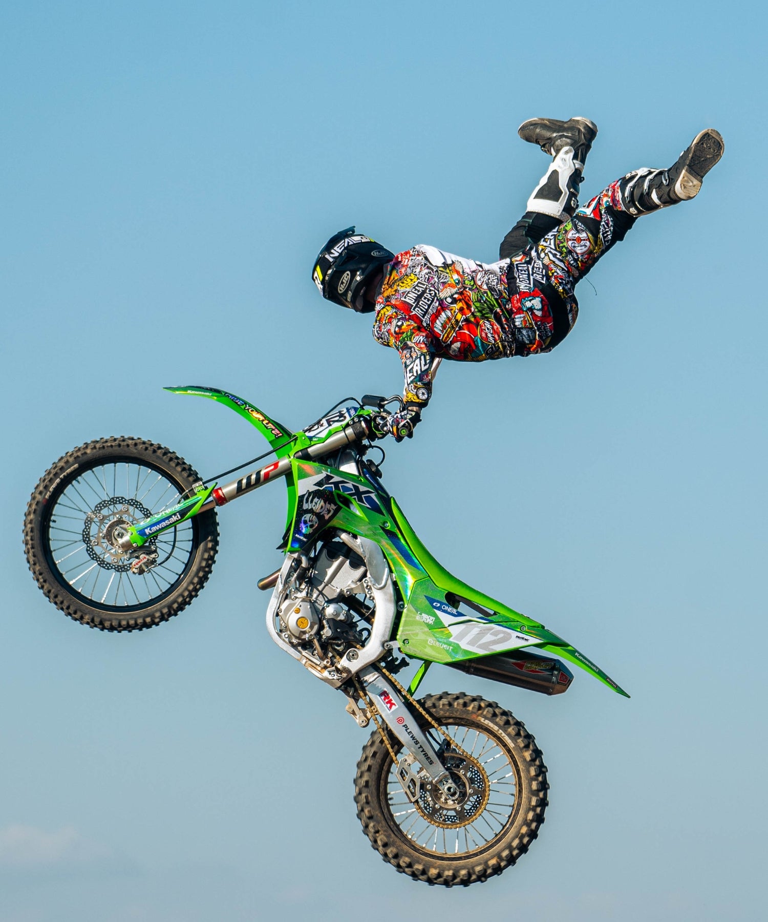 Ein Motocross-Fahrer in farbenfroher Ausrüstung führt einen Stunt in der Luft aus, indem er mit einer Hand den Lenker eines grünen Dirtbikes umklammert und dabei seine Beine und seinen Körper horizontal vor einem klaren blauen Himmel ausstreckt.