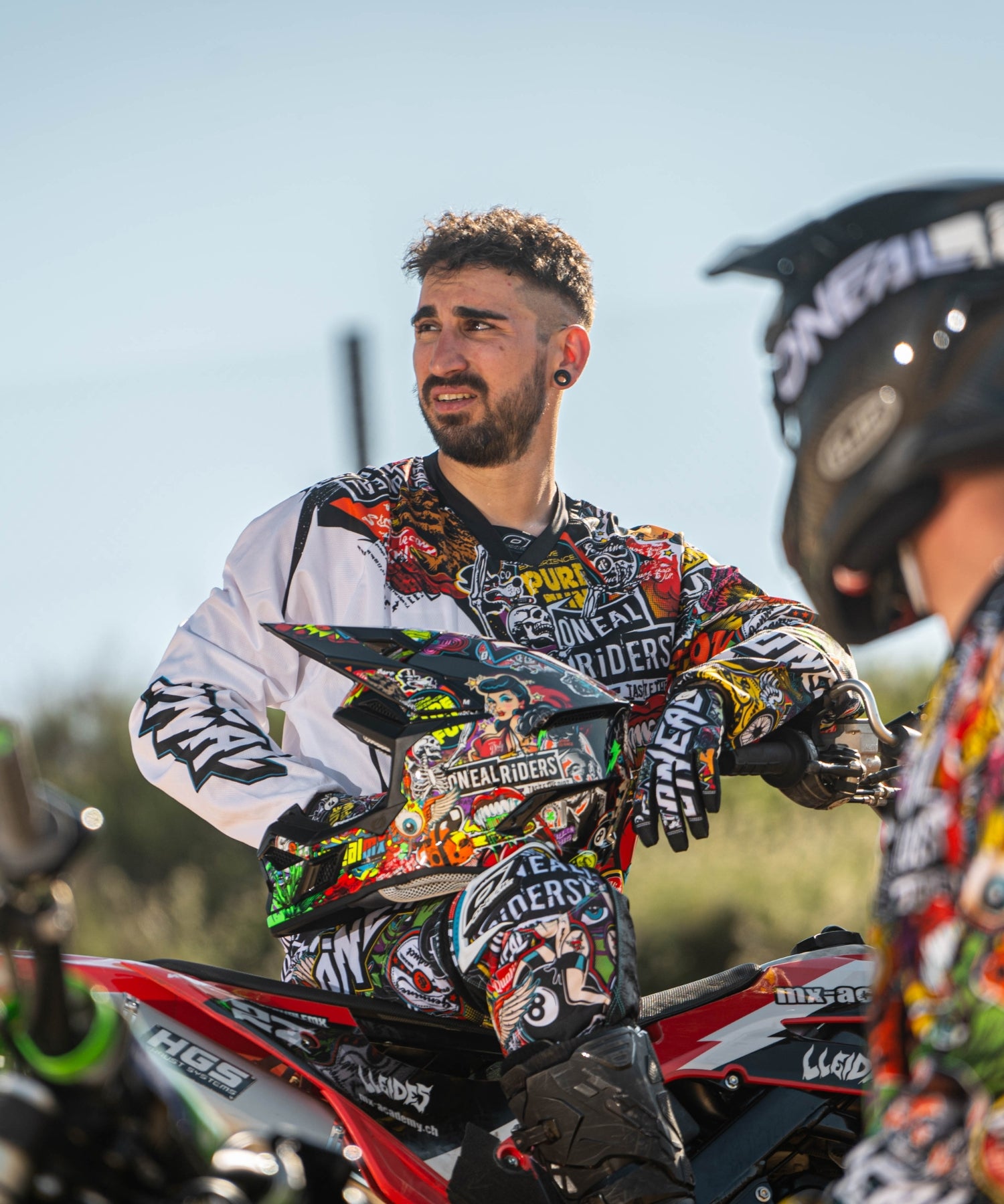 Ein Mann in farbenfroher, grafischer Motocross-Ausrüstung sitzt im Freien auf einem Dirtbike und blickt zur Seite, während im Vordergrund teilweise ein anderer Fahrer in ähnlicher Ausrüstung zu sehen ist.