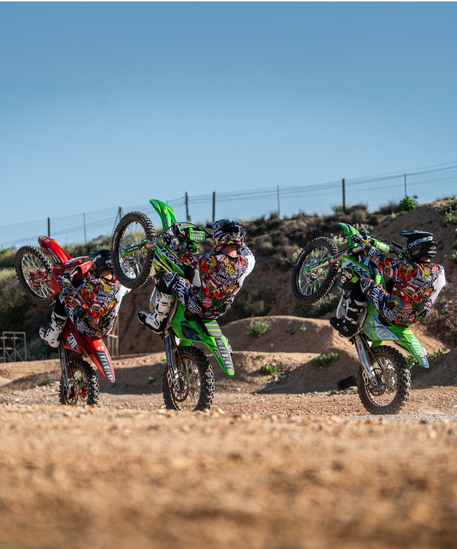 Drei Motocross-Fahrer in farbenfroher Ausrüstung machen auf ihren Dirtbikes Wheelies auf einer unbefestigten Strecke, mit einem klaren blauen Himmel und einem Zaun im Hintergrund.