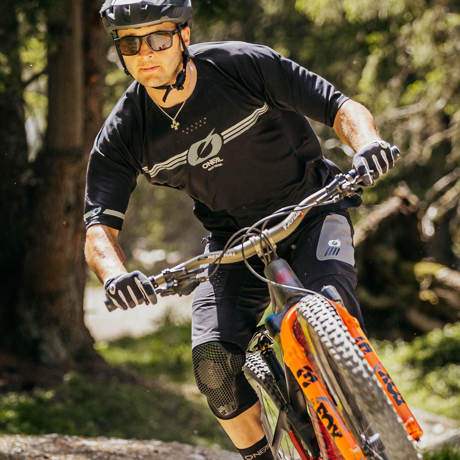 Eine Person mit schwarzem Helm, Sonnenbrille und schwarzer Radsportbekleidung mit Ellbogen- und Knieschützern fährt mit einem Mountainbike auf einem Waldweg. Das Fahrrad hat eine orangefarbene Vorderradgabel. Im Hintergrund sind Bäume und Grünflächen zu sehen.
