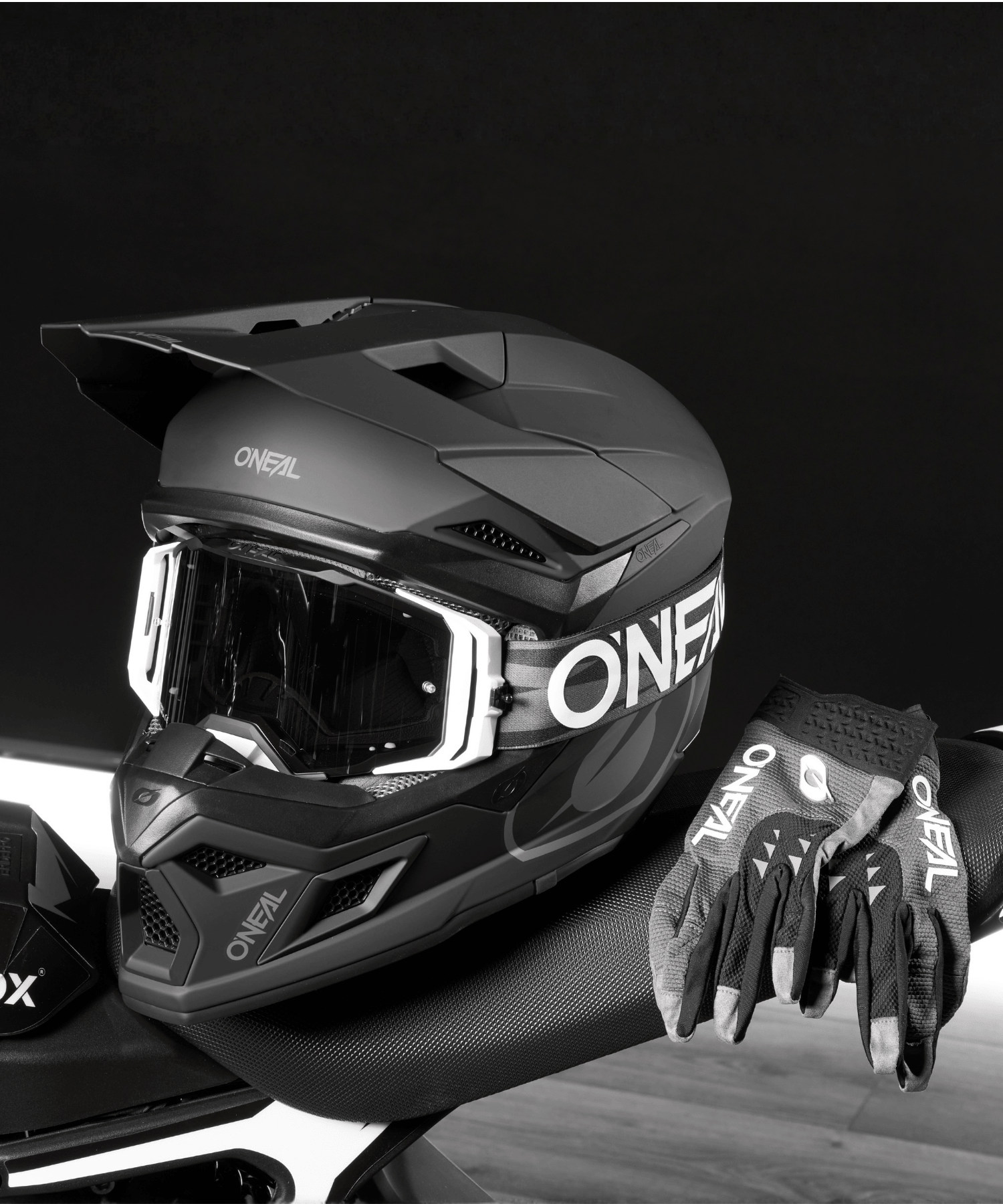 Ein schwarzer O'NEAL-Motocross-Helm mit Brille und ein Paar passender Handschuhe liegen auf dem Sitz eines Motorrads vor einem dunklen Hintergrund.
