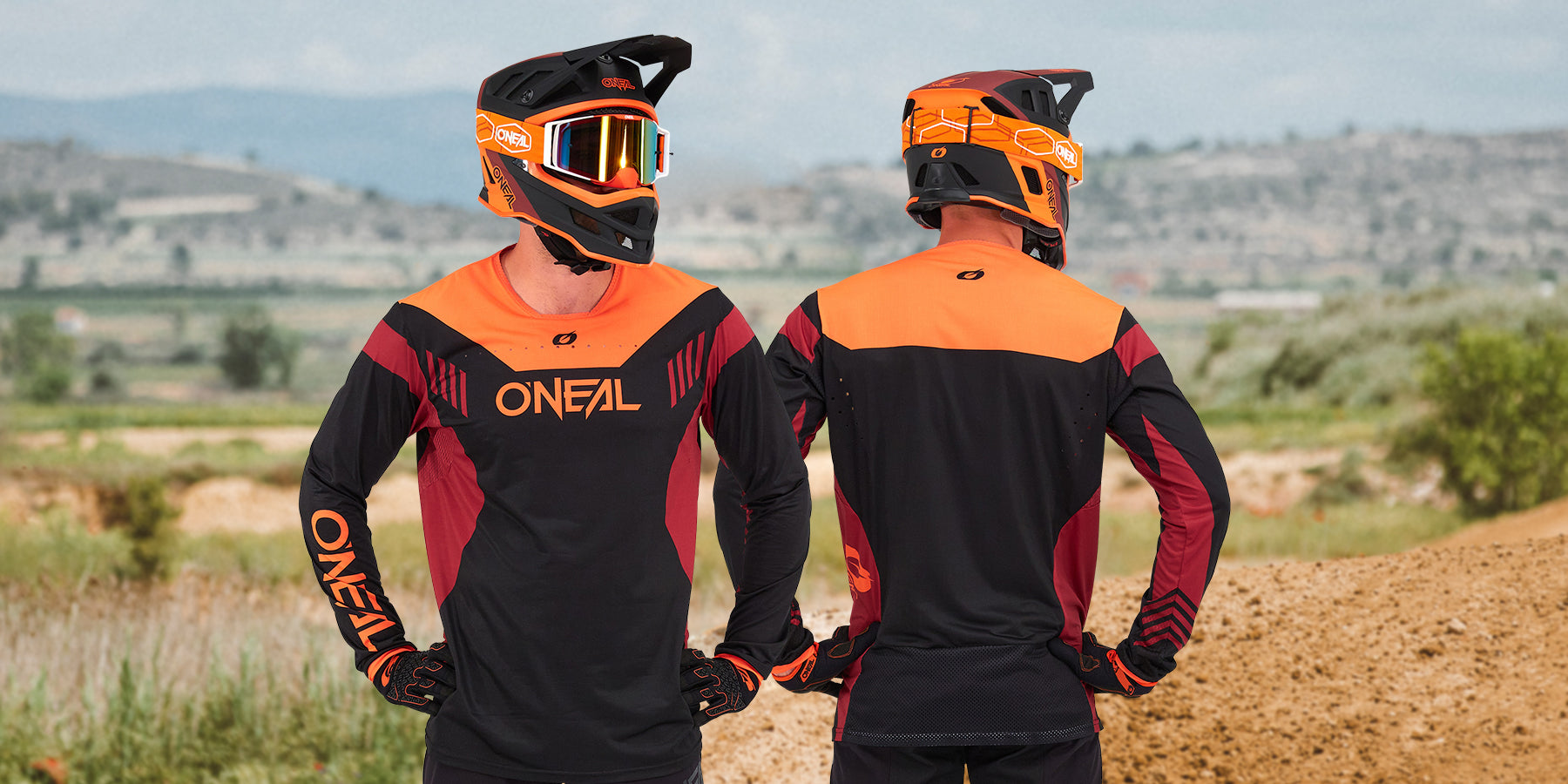 Zwei Motocrossfahrer in schwarzer, orangefarbener und roter O’Neal-Ausrüstung stehen auf einer unbefestigten Strecke. Der Fahrer links blickt nach vorne, während der Fahrer rechts wegblickt, sodass die Rückseite ihrer Uniformen zu sehen ist. Im Hintergrund ist eine hügelige Landschaft zu sehen.