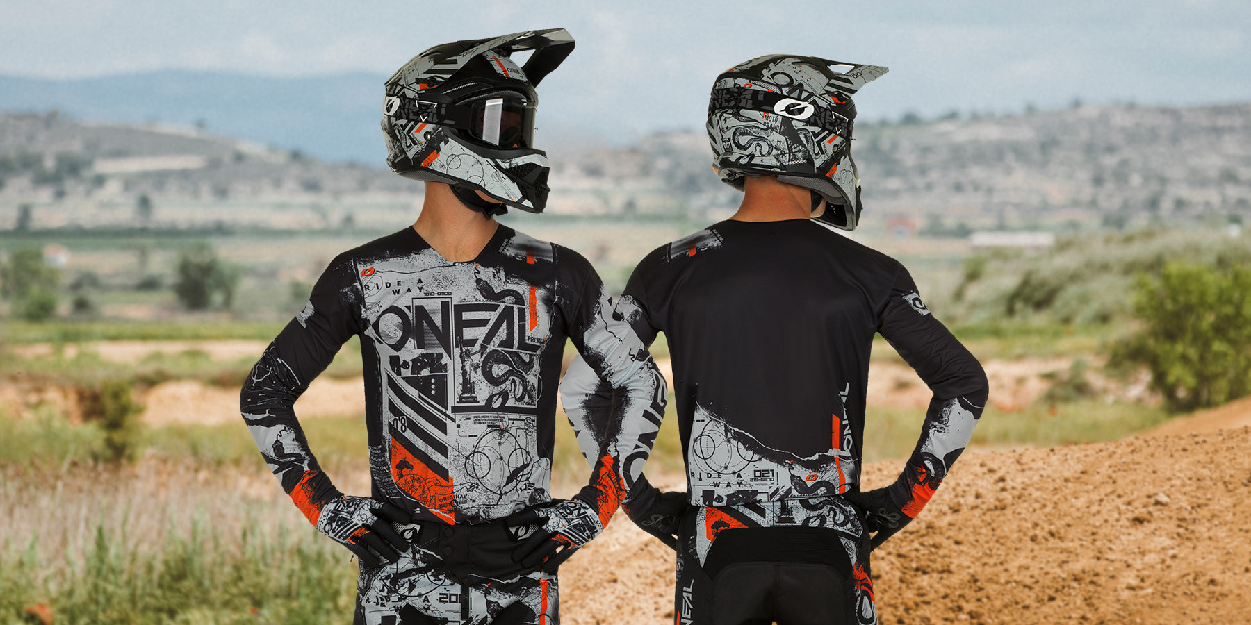 Zwei Motocrossfahrer in voller Motocross-Ausrüstung mit Helmen, Schutzbrillen, Handschuhen und gemusterten Anzügen stehen draußen auf einer unbefestigten Strecke. Einer blickt in die Kamera, der andere hat ihr den Rücken zugewandt. Die Landschaft im Hintergrund umfasst Felder und entfernte Hügel.