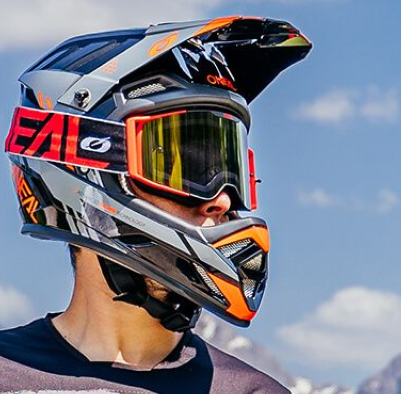 Eine Person trägt einen Integralhelm für Motorräder mit einem schwarz-grau-orangefarbenen Design. Der Helm verfügt über ein Visier und eine Schutzbrille mit gelber Tönung. Der Hintergrund zeigt einen klaren Himmel mit ein paar Wolken und einem Berggipfel in der Ferne.