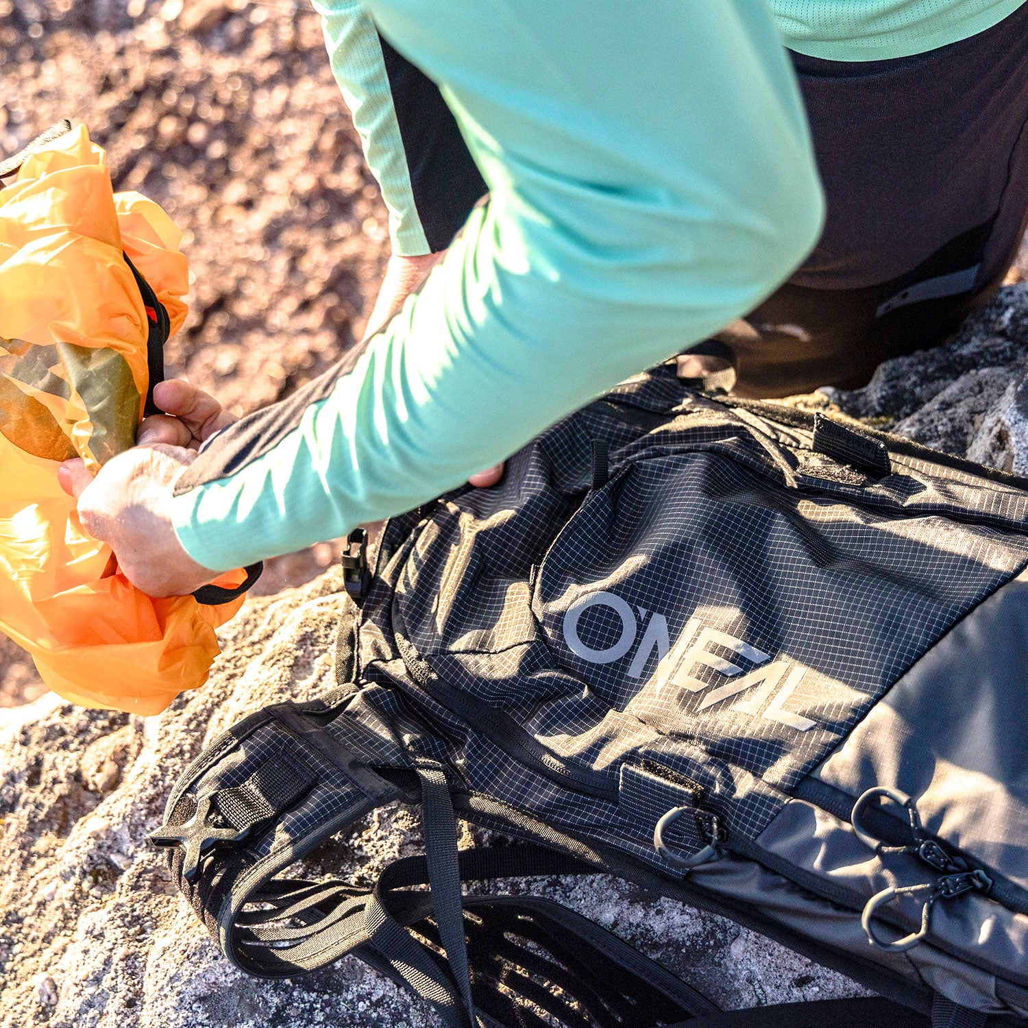 Eine Person mit einem hellgrünen langärmeligen Hemd packt eine orangefarbene Tasche neben einen schwarzen O'NEAL-Rucksack, der auf einer felsigen Oberfläche im Freien steht.