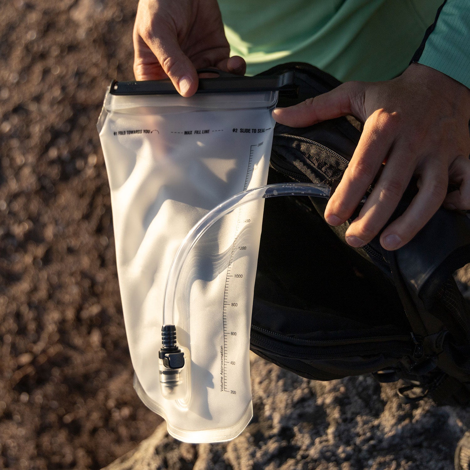 Eine Person hält ein durchsichtiges Trinkgefäß mit einem Trinkschlauch neben einen offenen Rucksack im Freien und bereitet sich darauf vor, das Gefäß darin zu verstauen.