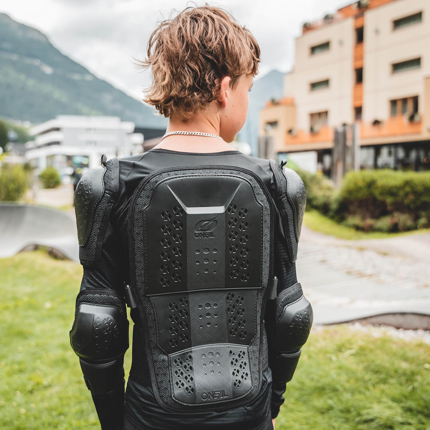 Eine Person mit lockigem Haar steht mit dem Rücken zur Kamera und trägt schwarze Schutzkleidung, darunter einen Rückenschutz und Ellbogenschützer. Sie befindet sich draußen auf einer Rasenfläche in der Nähe einiger moderner Gebäude. Im Hintergrund sind Berge zu sehen.