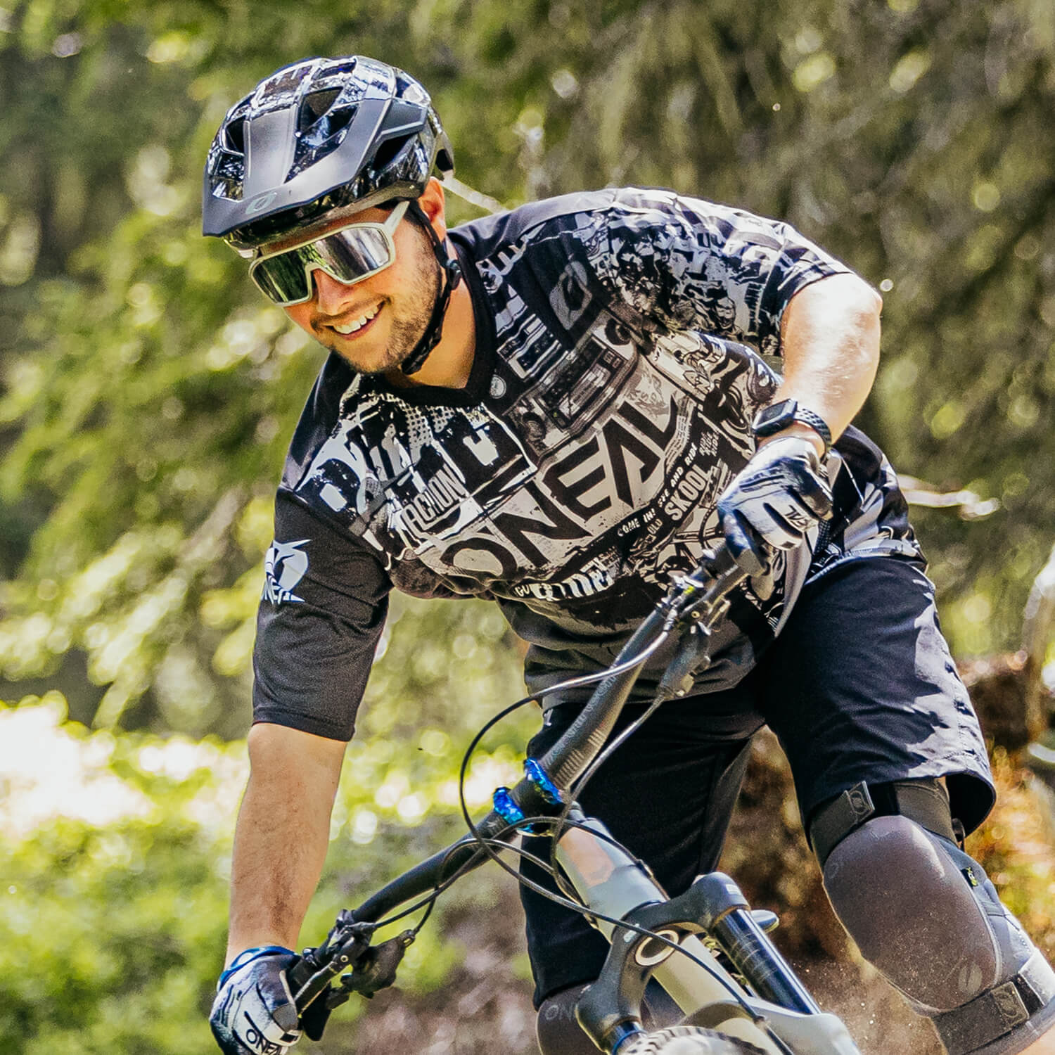 Ein Radfahrer mit schwarz-weißem Helm, Sonnenbrille und Schutzausrüstung fährt mit seinem Mountainbike durch ein Waldgebiet. Er lächelt, lehnt sich nach vorne und blickt konzentriert auf den vor ihm liegenden Weg.