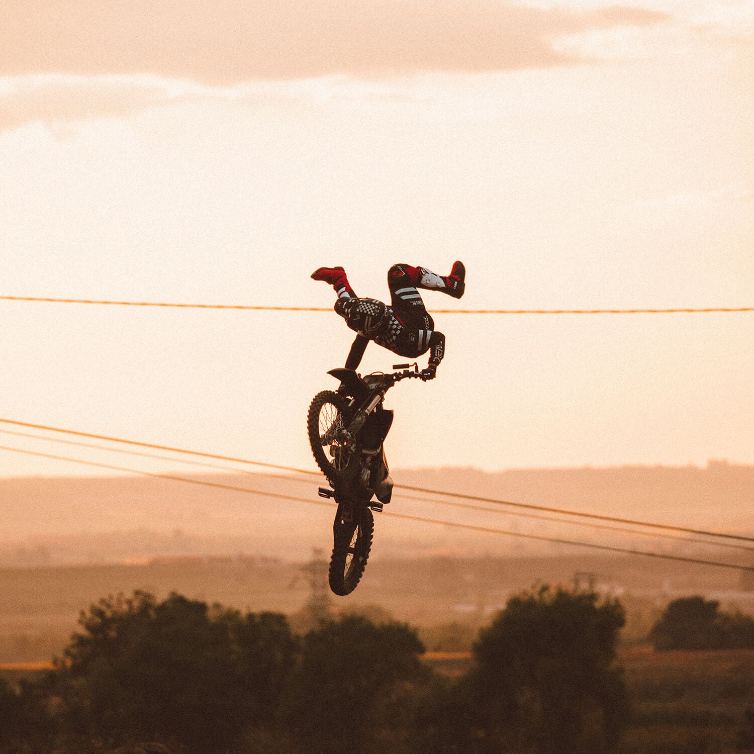 Ein Motorradfahrer vollführt einen waghalsigen Stunt, indem er bei Sonnenuntergang kopfüber über seinem Motorrad hängt. Die Beine des Fahrers strecken sich nach oben, während er sich an den Lenkern festhält, mit einer malerischen Landschaft im Hintergrund. Das Bild fängt einen Moment extremer Sport-Action ein.