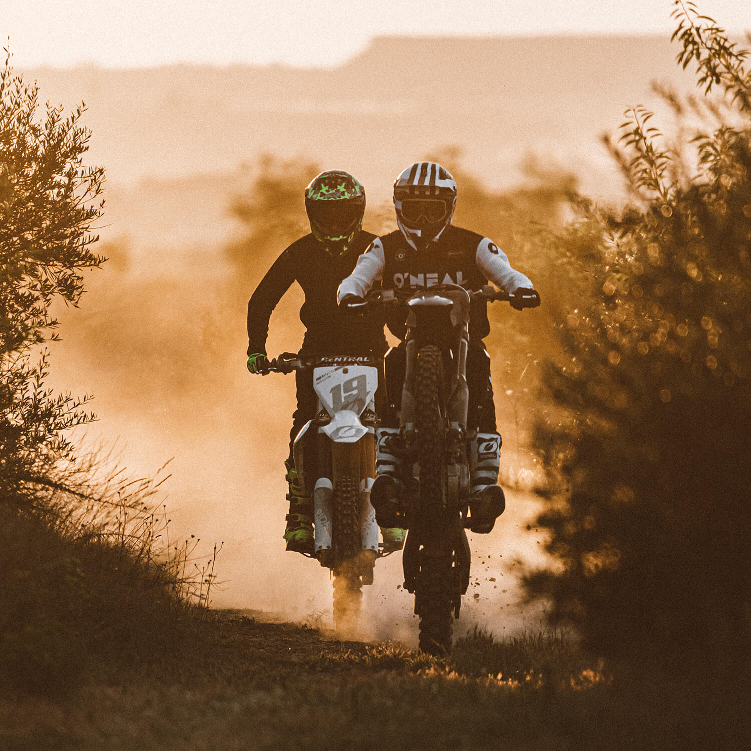 Zwei Motocrossfahrer, mit Helmen und Ausrüstung bekleidet, fahren bei Sonnenuntergang mit ihren Dirtbikes auf einem staubigen Pfad. Das warme, goldene Licht wirft lange Schatten und Silhouetten und schafft so eine dramatische Atmosphäre. Bäume und Gebüsch rahmen die Szene ein, während die Fahrer vorwärtsfahren.