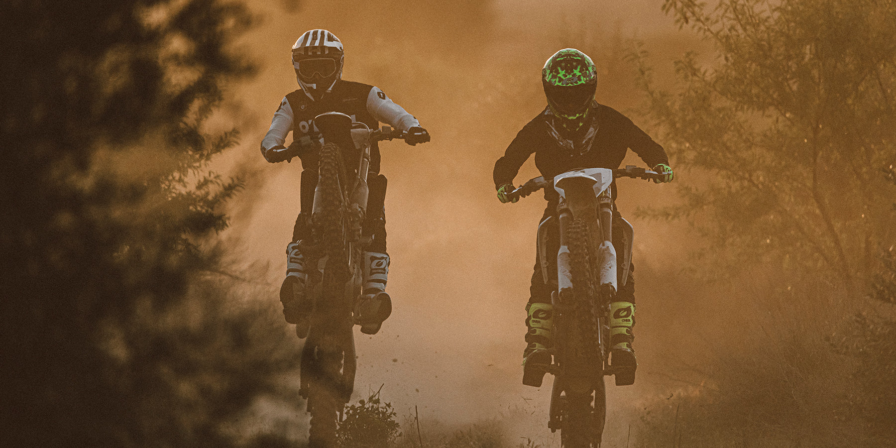Zwei Motocrossfahrer mit Helmen und Schutzausrüstung sind auf ihren Dirtbikes in der Luft und rasen bei Sonnenuntergang durch einen staubigen, von Bäumen gesäumten Pfad. Hinter ihnen steigen Staubwolken auf, die die Szene noch dramatischer ausleuchten.