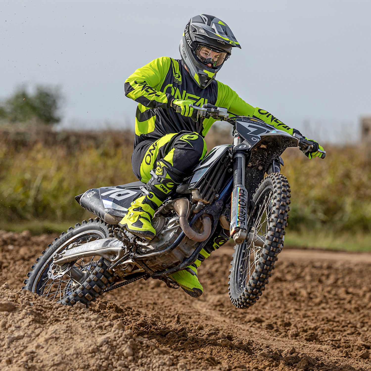Eine Person in neongrüner und schwarzer Motocross-Ausrüstung fährt mit einem Dirtbike über eine schlammige Strecke. Der Fahrer ist in der Luft, mit beiden Rädern vom Boden abgehoben und blickt nach vorne. Im Hintergrund sind verschwommenes Grün und ein bewölkter Himmel zu sehen.