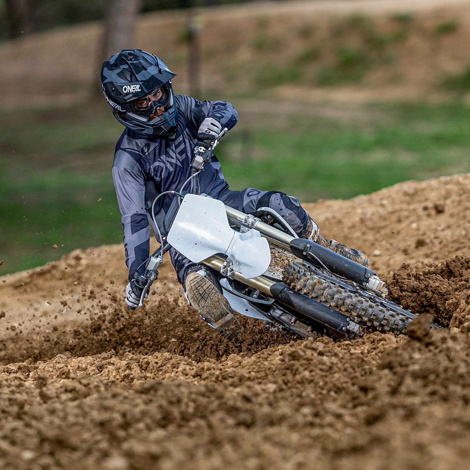 Eine Person in voller Motocross-Ausrüstung und mit Helm fährt mit einem Dirtbike über eine Sandpiste, wobei sie sich stark zur Seite neigt und dabei Sand und Dreck aufwirbelt. Der Hintergrund zeigt verschwommenes Grün und lässt an eine Offroad-Umgebung im Freien denken.