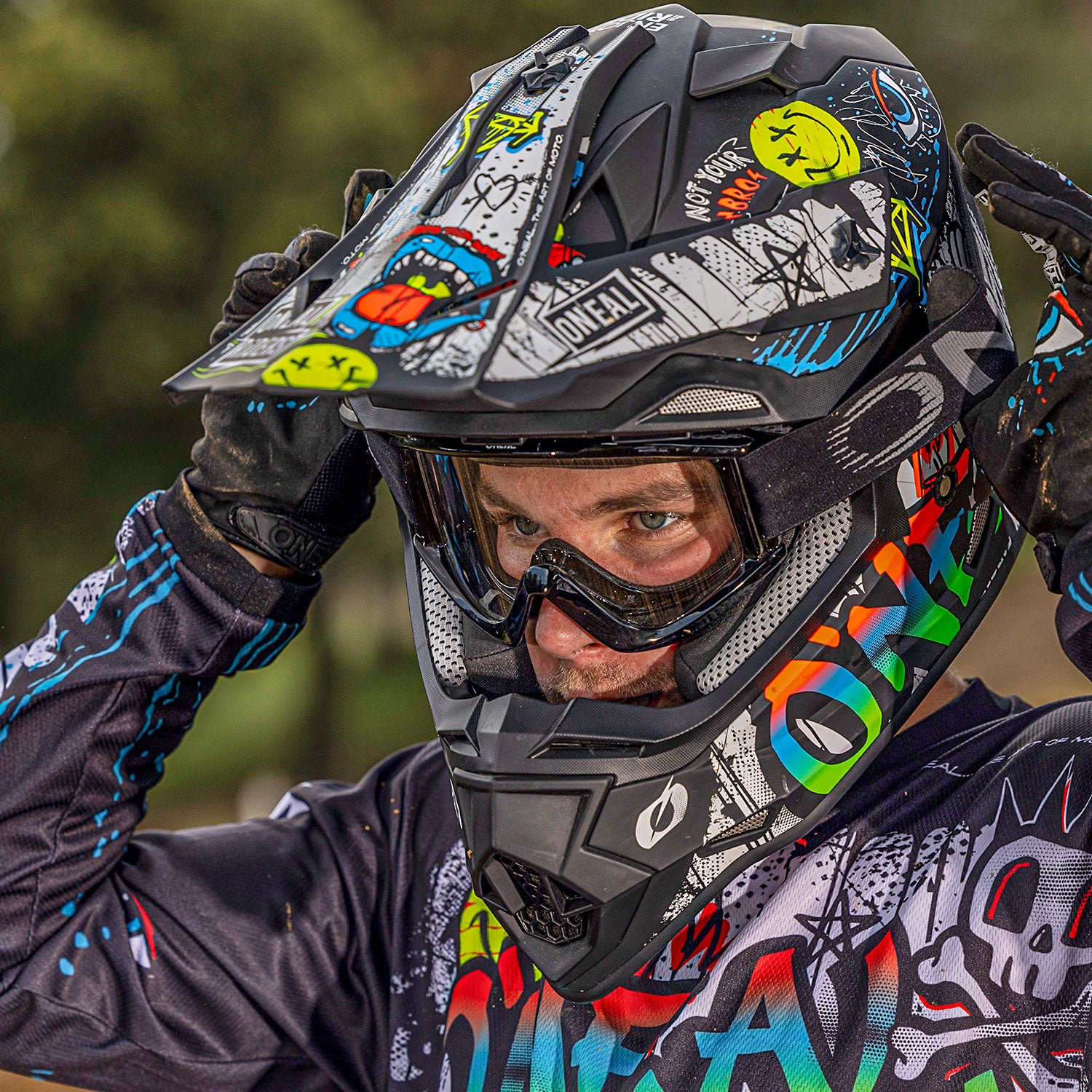 Eine Person trägt farbenfrohe Motocross-Ausrüstung, darunter einen Helm mit Graffiti-ähnlichen Mustern und eine Schutzbrille. Sie rückt den Helm zurecht, wobei sie beide Hände auf beiden Seiten ihres Kopfes hält. Der Hintergrund ist leicht verschwommenes Grün.
