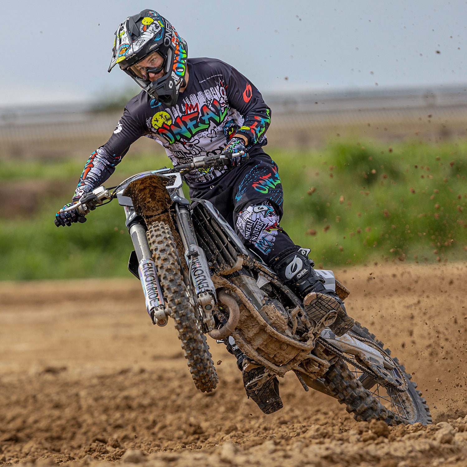 Ein Motocrossfahrer in schwarzem Outfit mit farbenfrohen Grafiken macht einen Wheelie auf einem Dirtbike. Im Hintergrund sind eine unbefestigte Strecke und verschwommene Vegetation zu sehen. Der Fahrer lehnt sich nach vorne, wobei Staub vom Hinterreifen spritzt. Dies fängt einen Moment voller Action und Können ein.