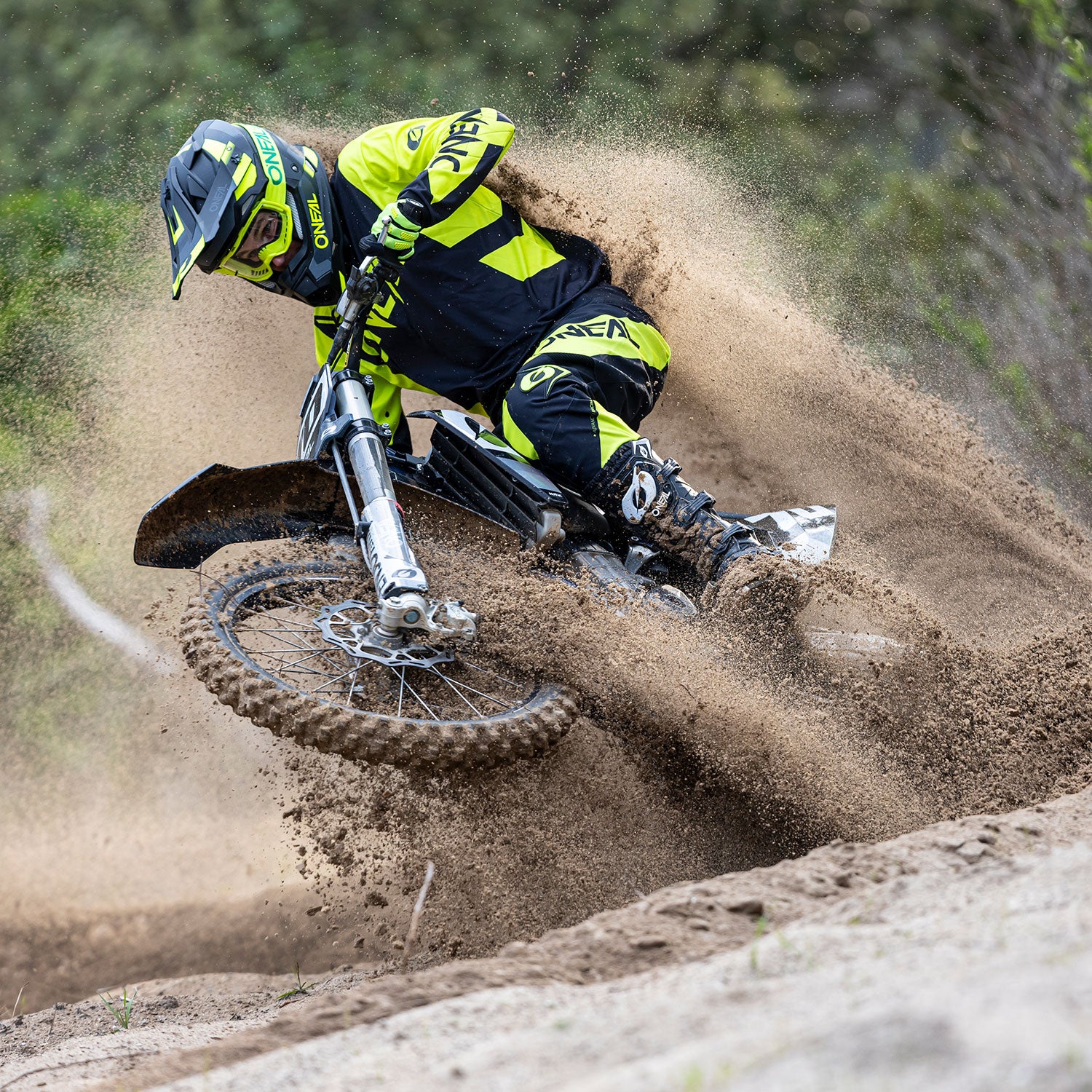 Ein Dirtbiker in neongelber und schwarzer Ausrüstung ist mitten in einer Kurve zu sehen, während er eine große Sandwolke aufwirbelt. Der Fahrer neigt sich scharf nach links und demonstriert Geschick und Kontrolle, während er durch das sandige Gelände navigiert. Der Hintergrund ist unscharf, was die Bewegung betont.
