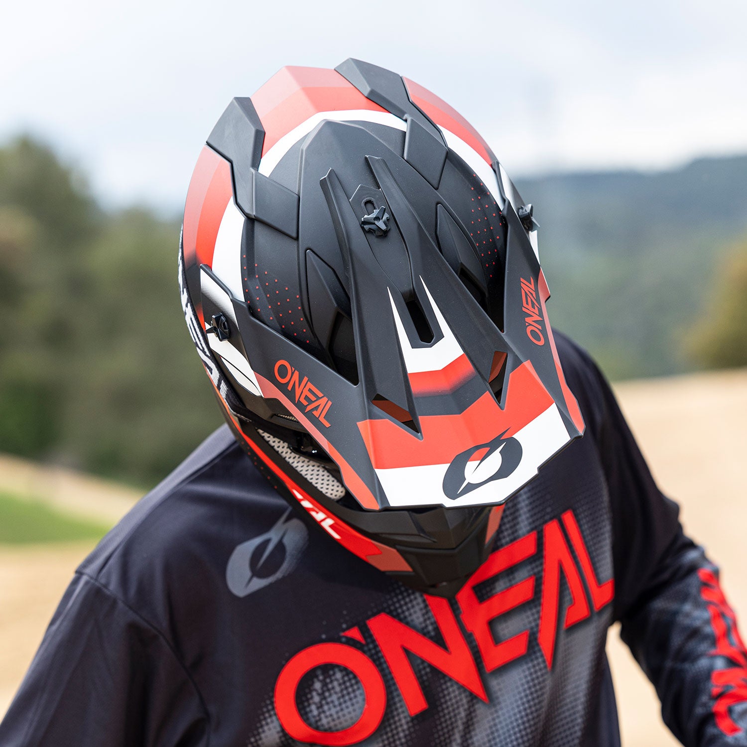 Eine Person mit einem schwarz-roten O'Neal-Motocrosshelm blickt nach unten und zeigt die Designdetails vorne und oben. Die Person trägt außerdem ein passendes schwarz-rotes O'Neal-Renntrikot. Der Hintergrund scheint eine natürliche Umgebung im Freien zu sein.