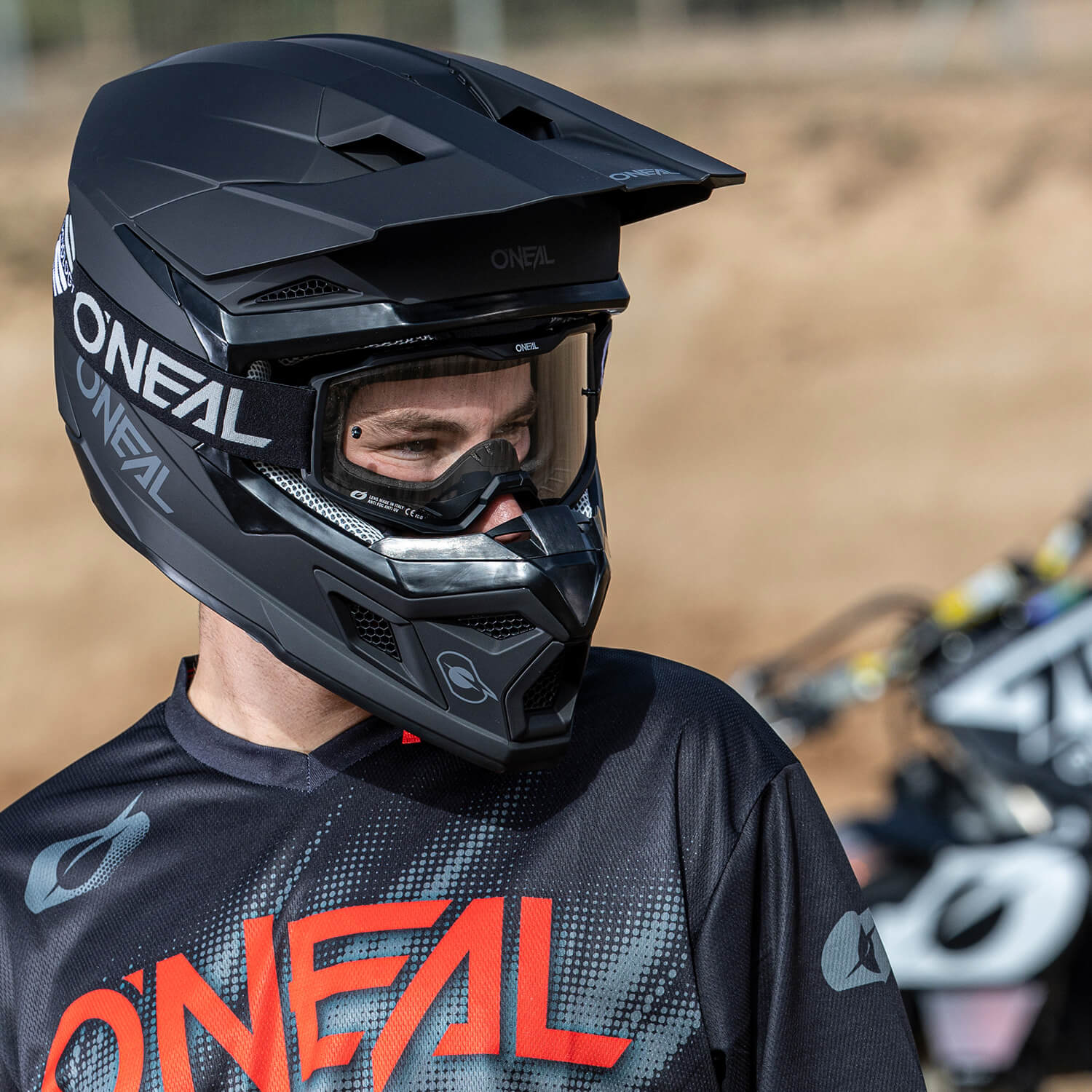 Eine Person mit einem schwarz-grauen O'Neal-Motocrosshelm und passender Ausrüstung steht im Freien, wahrscheinlich auf einer Motocrossstrecke. Sie blickt zur Seite, im Hintergrund sind Motorräder zu sehen.