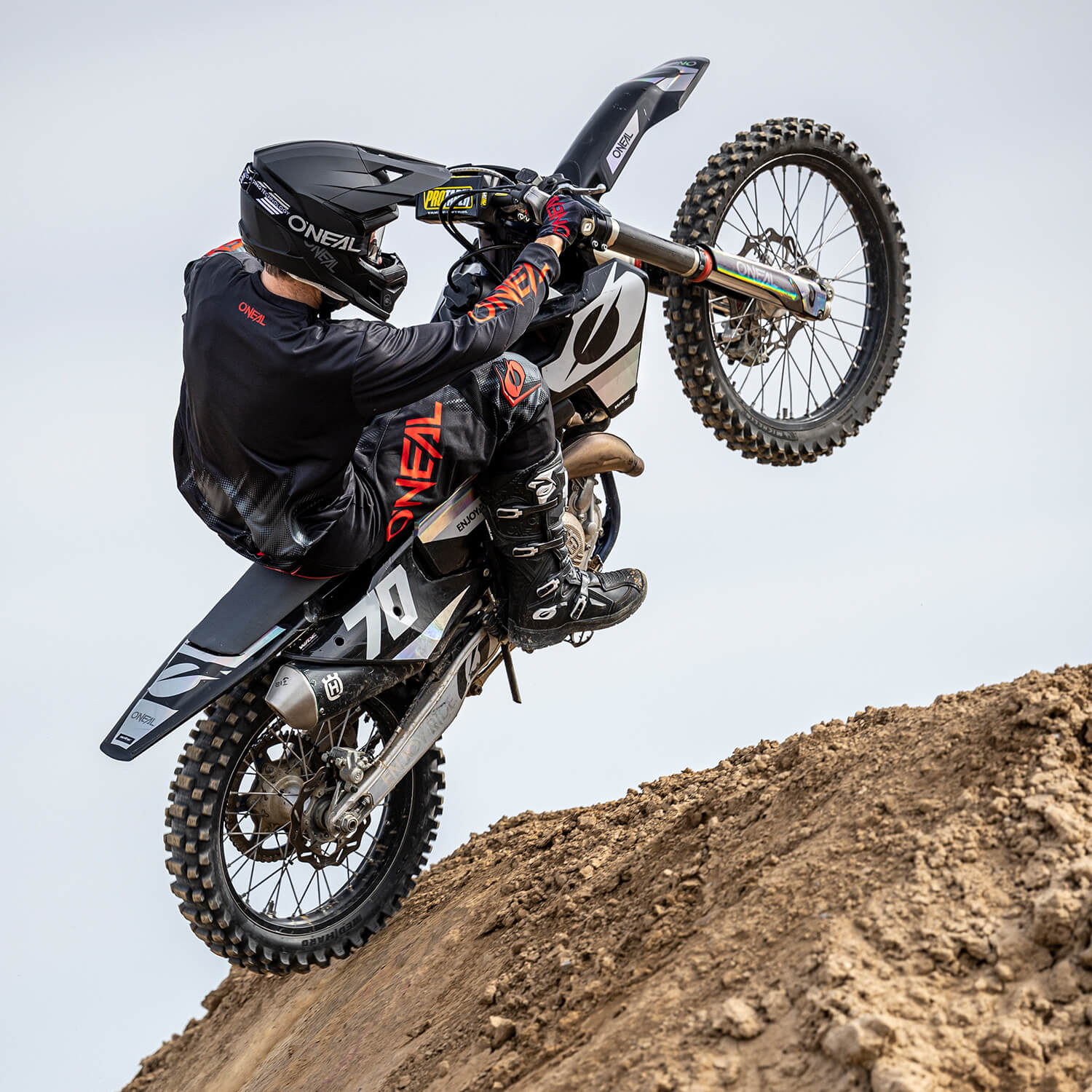 Ein Motocrossfahrer, der einen schwarzen O'Neal-Anzug und Helm trägt, macht einen Wheelie auf einem Erdhügel. Der Fahrer sitzt auf einem schwarz-weißen Dirtbike, auf dessen Vorderseite die Nummer 70 zu sehen ist. Der Himmel ist klar und hellgrau.