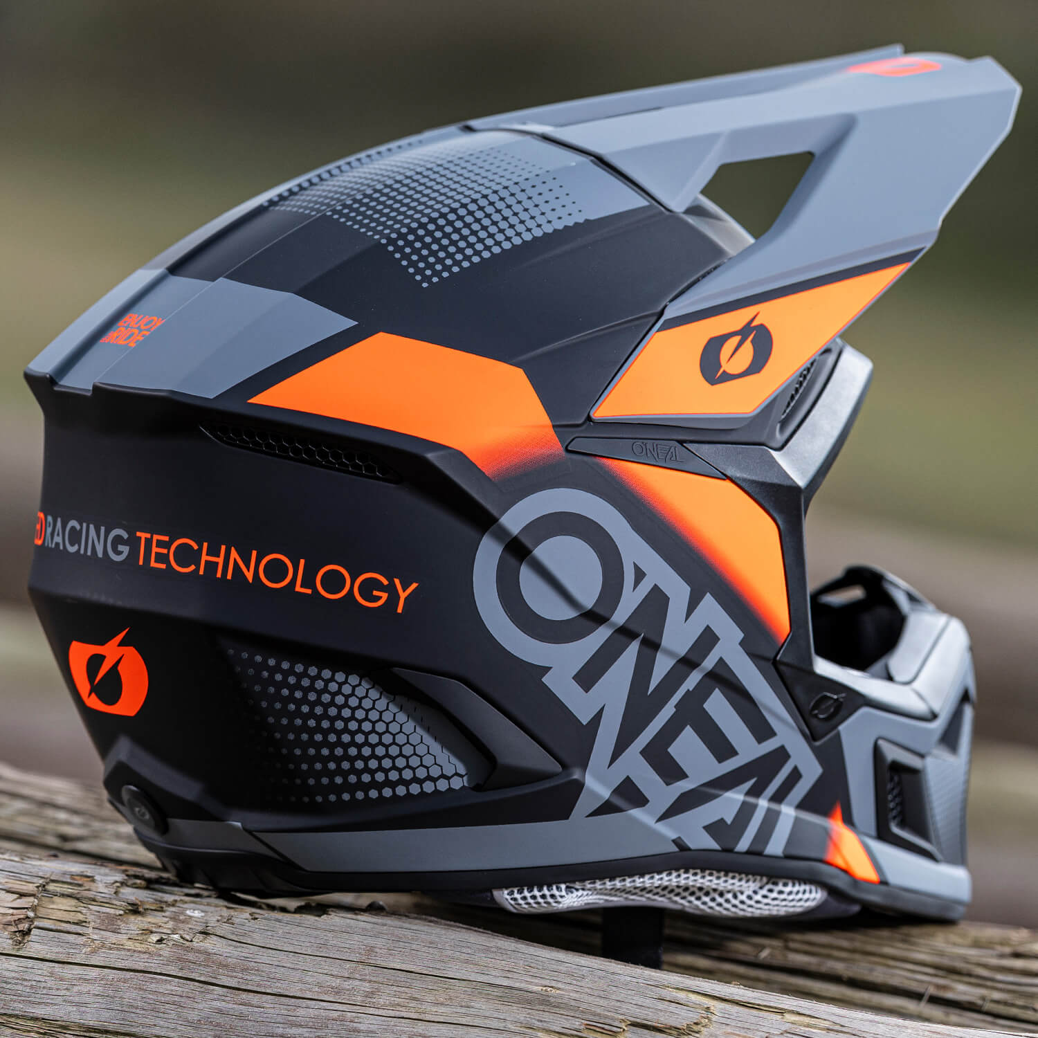 Eine Nahaufnahme eines eleganten, modernen Motorradhelms, der auf einer Holzoberfläche ruht. Der Helm ist überwiegend schwarz mit kräftigen orangefarbenen Akzenten und trägt die Worte „Racing Technology“ und „ONEAL“ in großer Schrift. Das Design ist eckig und futuristisch.