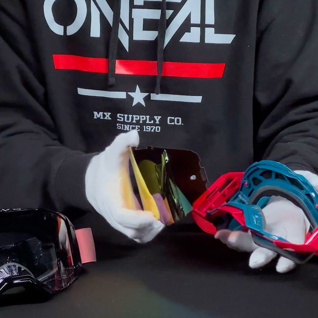 Eine Person mit weißen Handschuhen und einem schwarzen Kapuzenpullover mit der Aufschrift „O’NEAL MX SUPPLY CO.“ wechselt die Linse einer Motocross-Brille, während eine andere Brille auf dem Tisch liegt.