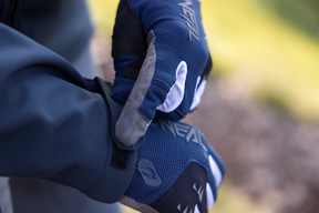 Eine Nahaufnahme einer Person, die marineblaue Softshell-Handschuhe O'NEAL CYCLONE trägt und den rechten mit der linken Hand zurechtrückt. Das atmungsaktive Material von O'NEAL sorgt für Komfort, während der Hintergrund unscharf ist, um diese wasserfesten Handschuhe hervorzuheben.