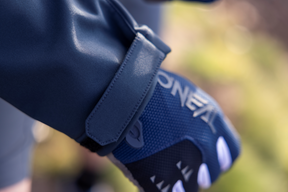 Nahaufnahme der Hand einer Person, die einen blau-schwarzen Handschuh mit weißen Akzenten trägt. Der wasserfeste Handschuh mit Klettverschluss und dem Markennamen „O'Neal“ passt gut zu ihrer dunklen O'NEAL CYCLONE Soft Shell-Jacke. Der Hintergrund ist unscharf.