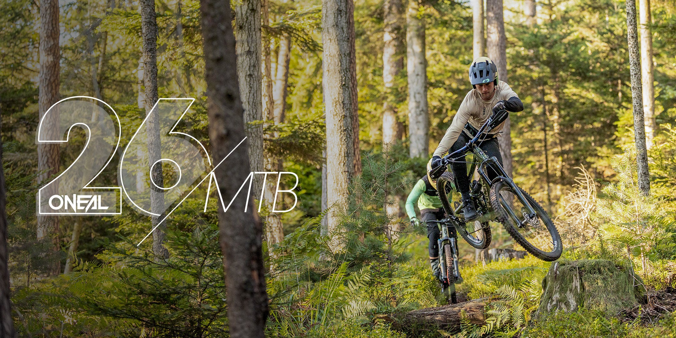 Ein Mountainbiker mit Helm und Ausrüstung springt auf einem Waldweg über einen Baumstamm; ein anderer Fahrer folgt ihm. Das Sonnenlicht fällt durch die hohen Bäume. Der Text auf der linken Seite lautet "26% O'NEAL MTB.