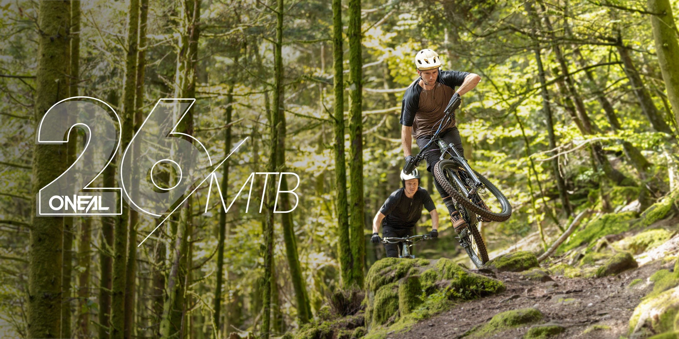 Zwei Mountainbiker fahren durch einen Waldweg. Der erste Fahrer hebt das Vorderrad über Felsen, der zweite folgt dicht dahinter. Sie sind von Bäumen und moosbewachsenem Boden umgeben. Der Text lautet "26 ONEAL MTB.