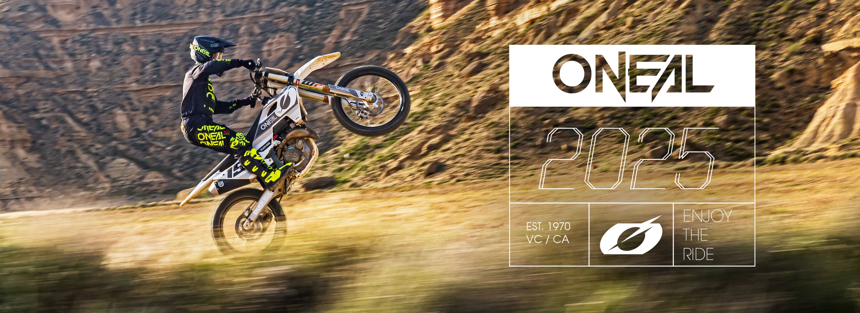 Ein Motocrossfahrer macht auf einem Dirtbike einen Wheelie in einer felsigen, bergigen Landschaft. Der Fahrer trägt schwarze und neongrüne Ausrüstung. Die Texte „ONEAL“ und „2025“ sind gut sichtbar, zusammen mit dem Gründungsjahr, dem Standort und dem Slogan „Enjoy the Ride“.