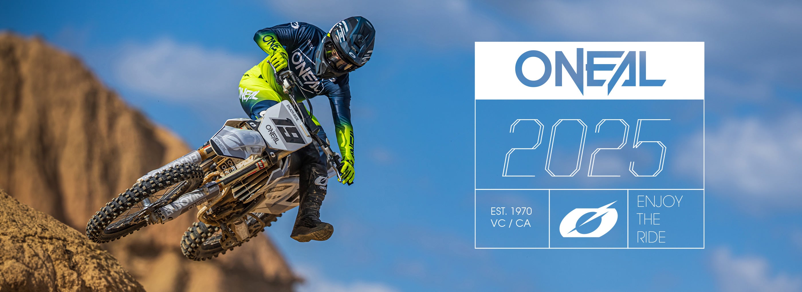 Ein Motocrossfahrer in voller Montur macht vor einer Kulisse aus zerklüftetem Gelände einen Sprung auf einem Dirtbike. Das Bild enthält den Text „O'NEAL 2025, EST. 1970 VC/CA, ENJOY THE RIDE“.